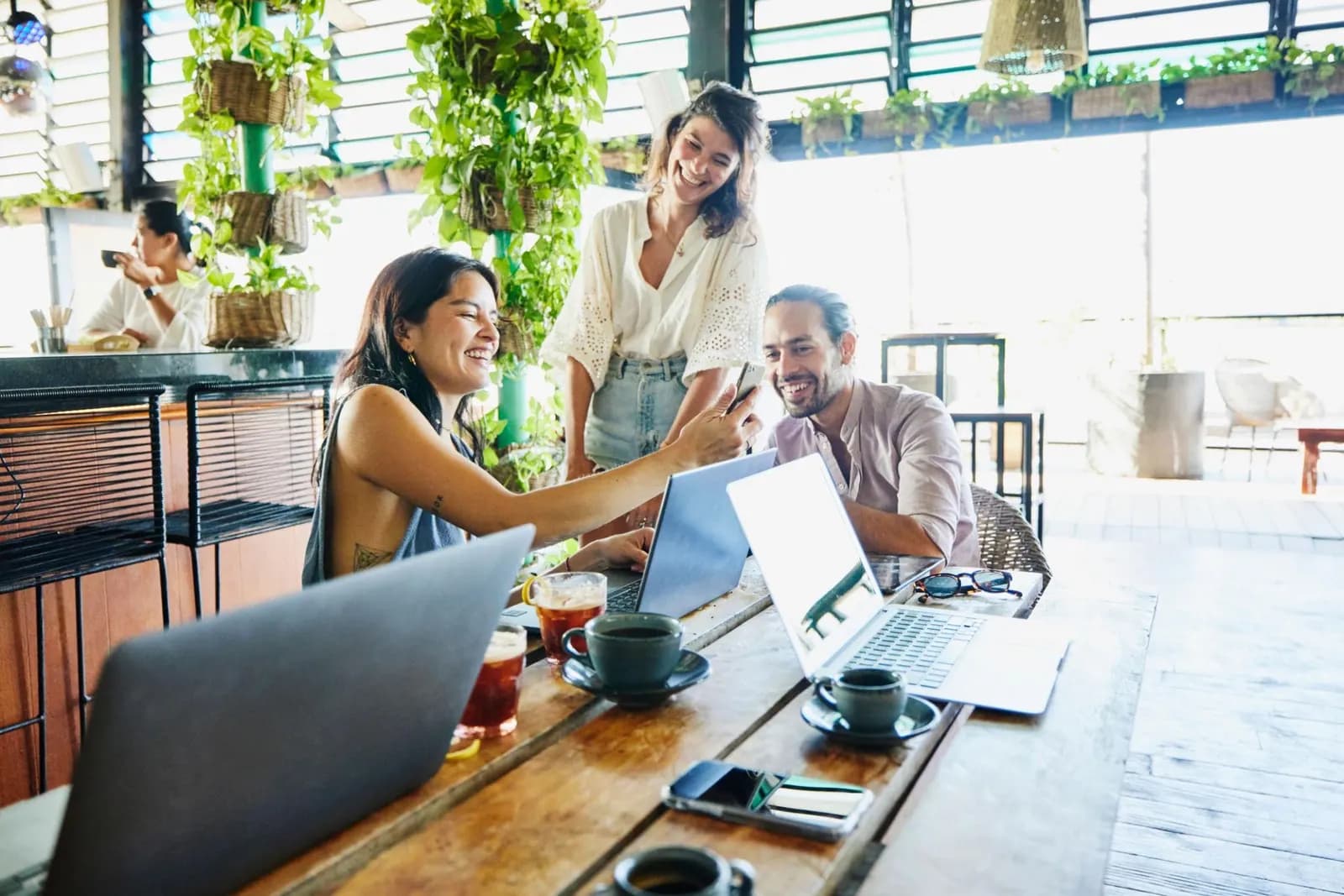 Three people laughing around laptops and coffee in a bright hostel common area with hanging plants